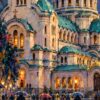 Детайл: осветени арки и зелени покриви на храма над фигури с чадъри на мокрия площад