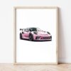Постер с розов Porsche GT3 RS в светла дървена рамка, облегнат на бяла стена върху паркет